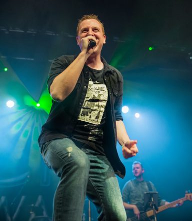 Singer performing energetically on stage, with a guitarist in the background and vibrant lighting.