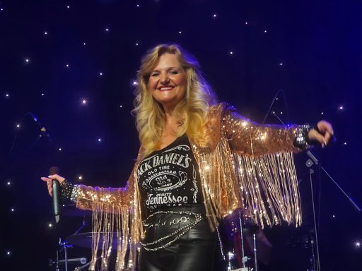 A smiling performer in a sparkling jacket sings on stage, with a light backdrop.