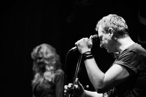 Man singing into a microphone with a woman slightly blurred in the background.