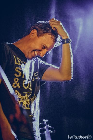 A man smiles while lightly touching his head, with stage lighting behind him.